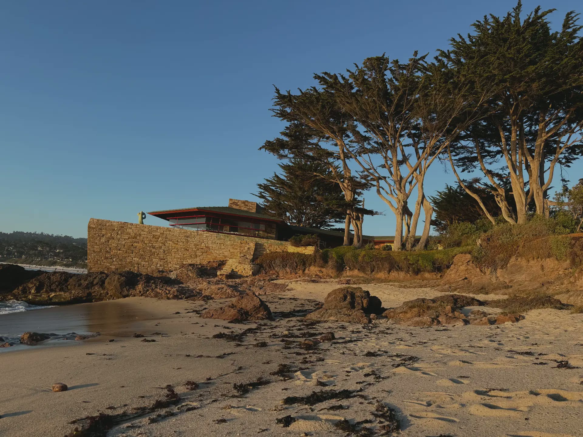 A stone-walled modernist house above the white sand and cypress of Carmel Beach at golden hour